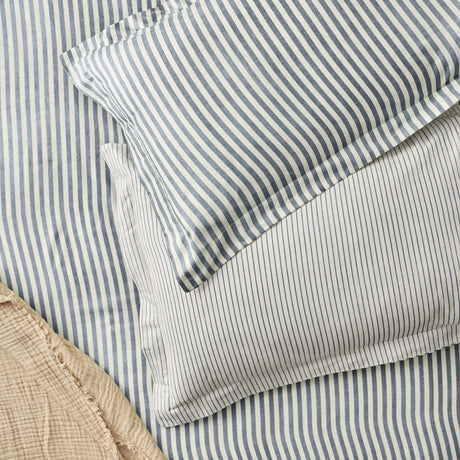 Hebden Melange Cotton Pillowcase Pair in Navy, showing slate blue heathered stripes on off-white cotton, layered on bedding with a plain beige textured throw nearby.