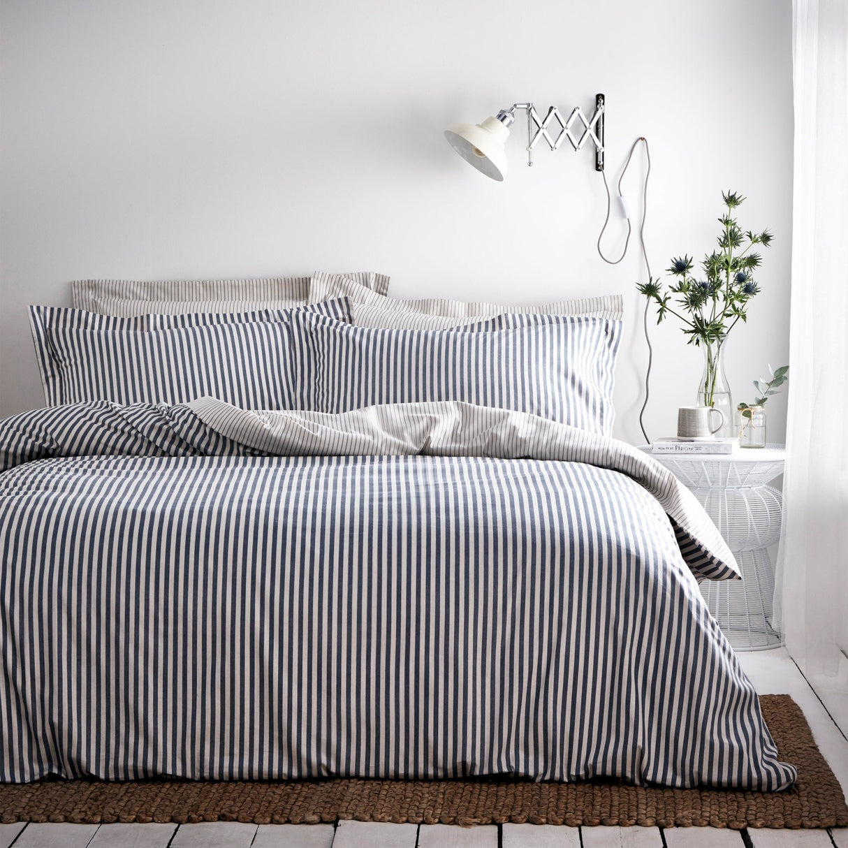Hebden Cotton Striped Duvet Cover Set in navy on a bedroom bed, with matching pillowcases, a white side table, wall lamp, and vase of flowers.
