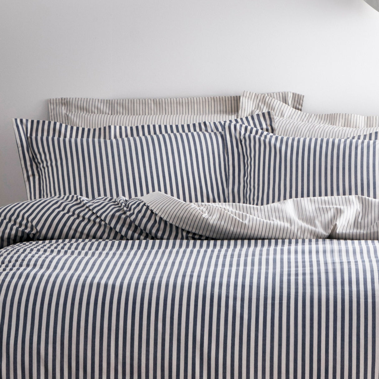 Hebden Cotton Striped Duvet Cover Set in Navy, dressed on a bed with matching pillowcases, showing a narrow navy and off-white stripe against a plain grey wall.