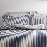 Hebden Cotton Striped Duvet Cover Set in Navy, dressed on a bed with matching pillowcases, showing a narrow navy and off-white stripe against a plain grey wall.
