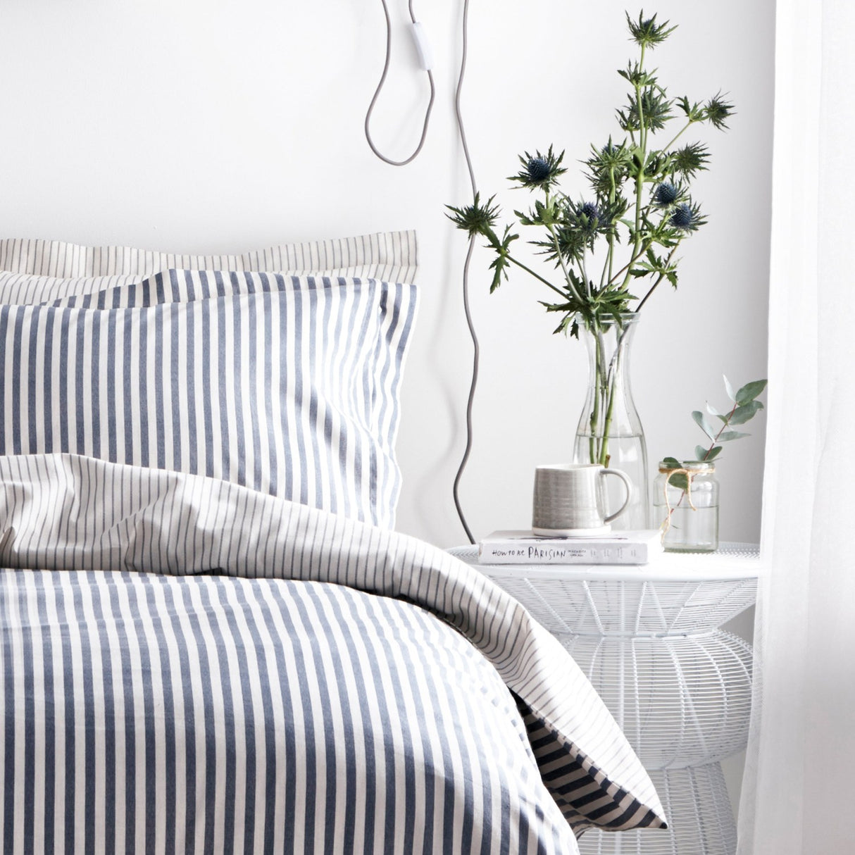 Hebden Cotton Striped Duvet Cover Set in Navy on a bedroom bed, with matching pillowcases, styled beside a white wire side table, mug, books, and a glass vase of thistle flowers.