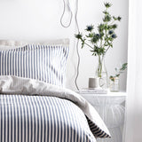 Hebden Cotton Striped Duvet Cover Set in Navy on a bedroom bed, with matching pillowcases, styled beside a white wire side table, mug, books, and a glass vase of thistle flowers.