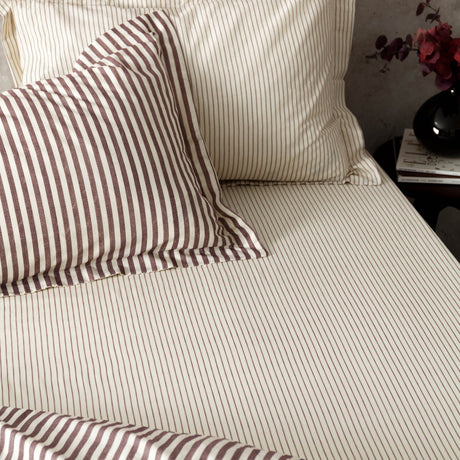 Hebden Striped Cotton Fitted Sheet in Beetroot, cream cotton bedding with fine red stripes, shown with matching striped pillowcases on a bed.