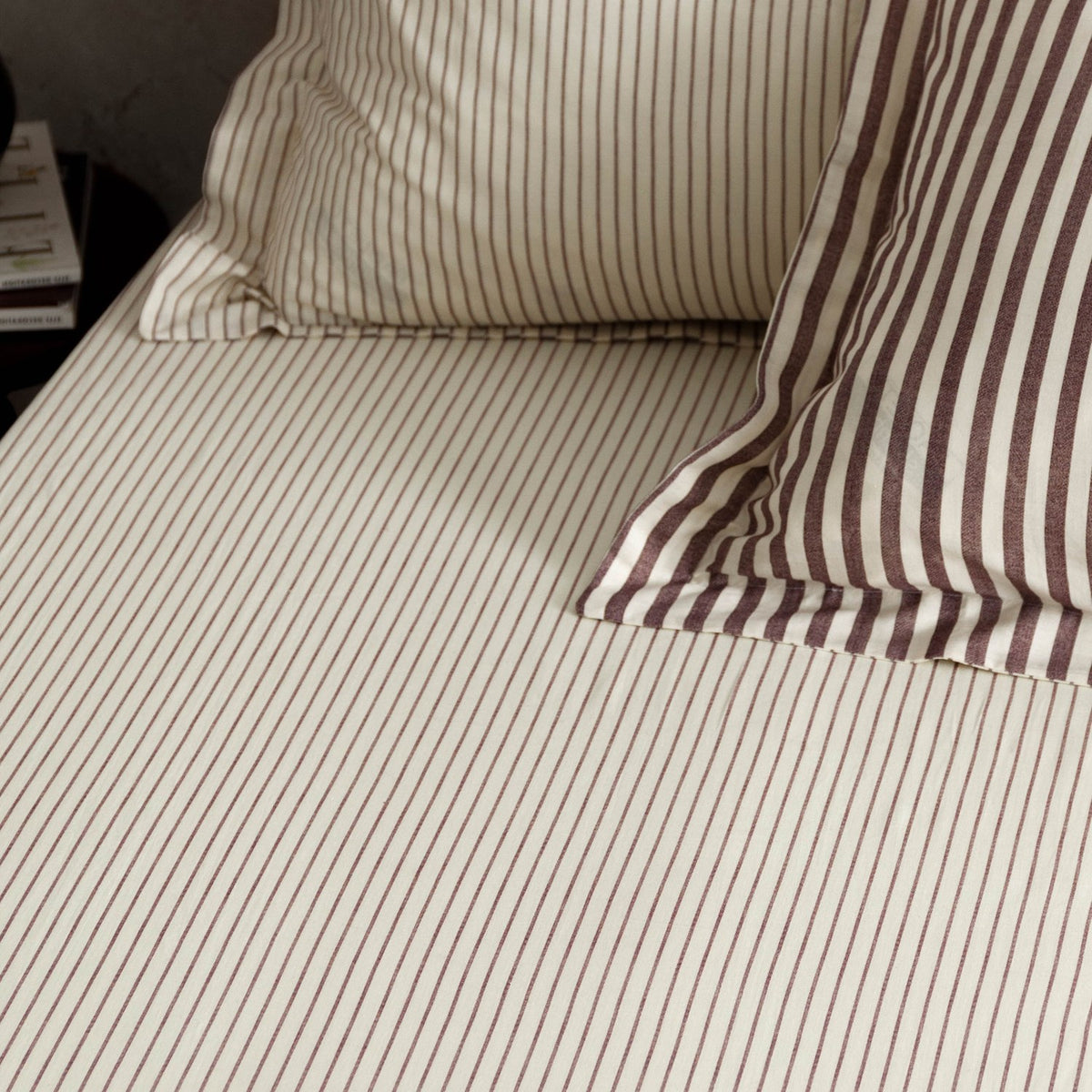 Hebden Striped Cotton Fitted Sheet in Beetroot, cream cotton with narrow muted red stripes, shown fitted to a bed with matching pillowcases.