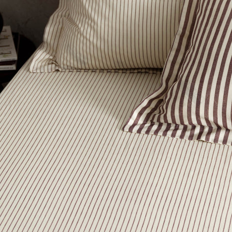 Hebden Striped Cotton Fitted Sheet in Beetroot, cream cotton with narrow muted red stripes, shown fitted to a bed with matching pillowcases.