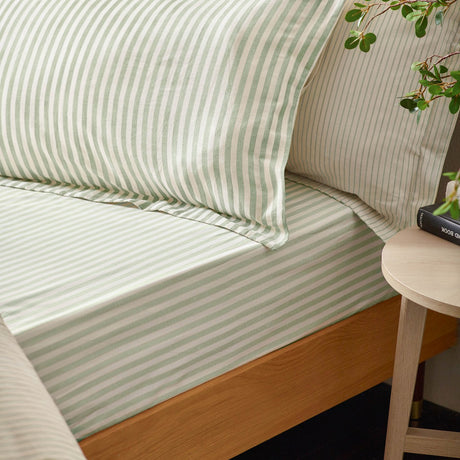 Hebden Reversible Cotton Fitted Sheet in pale green stripe on cream ground, shown fitted on a wooden bed with matching striped pillowcases in a bedroom setting.