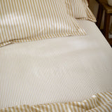 Hebden Striped Cotton Fitted Sheet in honey yellow, dressed on a bed with matching pillowcases, showing a cream ground with a narrow sandy gold stripe.