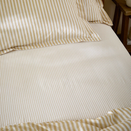 Hebden Striped Cotton Fitted Sheet in honey yellow, dressed on a bed with matching pillowcases, showing a cream ground with a narrow sandy gold stripe.