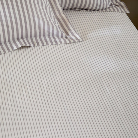 Hebden Striped Cotton Fitted Sheet in mauve-grey, made from cotton with a narrow striped pattern, shown on a bed with matching pillowcases.
