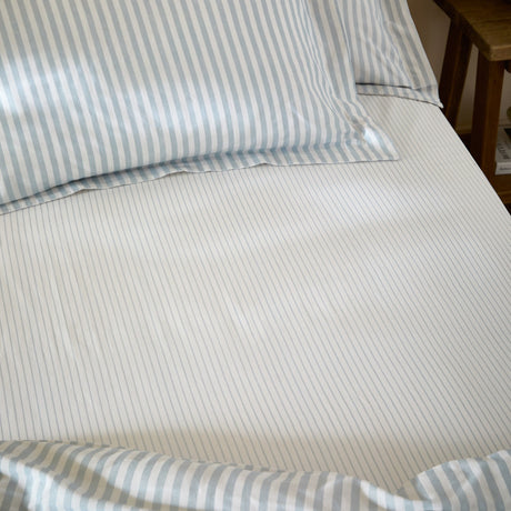 Hebden Striped Cotton Fitted Sheet in Mineral Blue on a made bed, showing narrow blue and white stripes on cotton fabric with matching striped pillowcases.