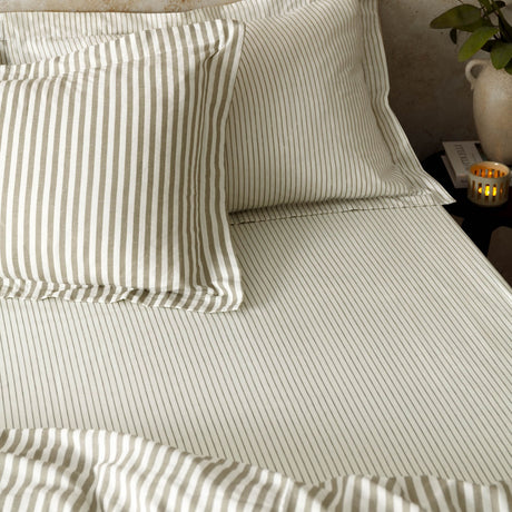 Hebden Striped Cotton Fitted Sheet in Mole, styled on a bed with matching pillowcases in cream and taupe-grey pinstripe cotton, set in a bedroom with a bedside candle and vase.