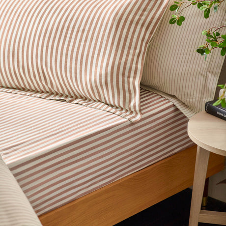 Hebden Cotton Fitted Sheet in cream with pecan and terracotta stripes, shown fitted on a wooden bed frame with matching striped pillowcases in a bedroom setting.