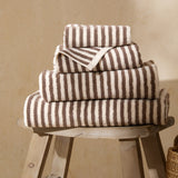 Hebden Striped Cotton Towels in chocolate brown and cream, four sizes stacked on a wooden stool showing the vertical stripe design.