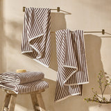 Hebden Striped Cotton Towels in chocolate brown and cream, hanging from brass rails and folded on a wooden stool in a bathroom setting.