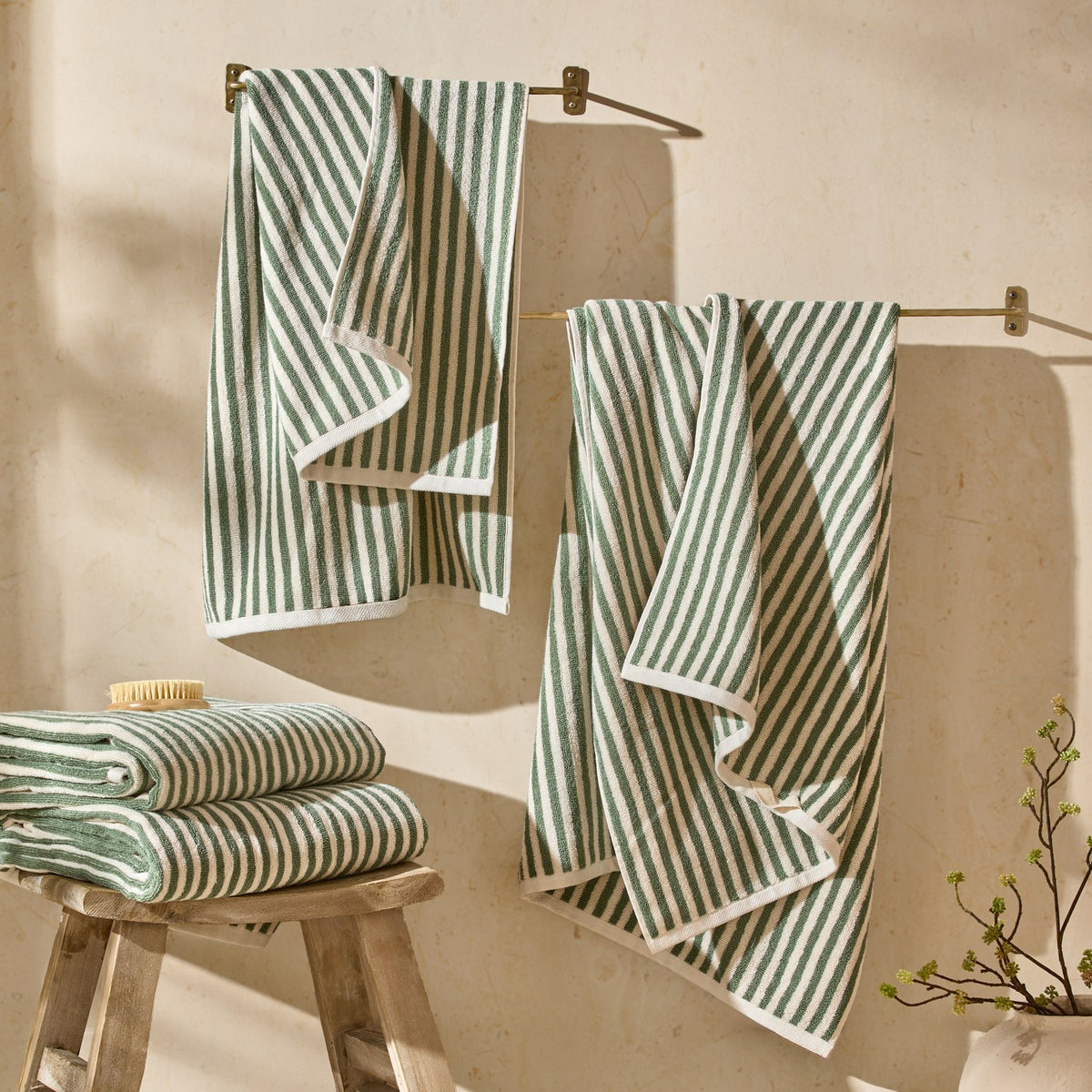 Hebden Striped Cotton Towels in Forest hanging from brass rails and folded on a wooden stool in a bathroom setting, showing green and cream vertical stripes with white trim.