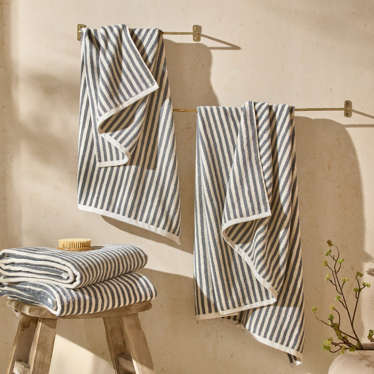 Hebden Striped Cotton Towels in grey and cream hang from brass rails and stack folded on a wooden stool in a bathroom setting.