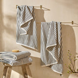 Hebden Striped Cotton Towels in grey and cream hang from brass rails and stack folded on a wooden stool in a bathroom setting.