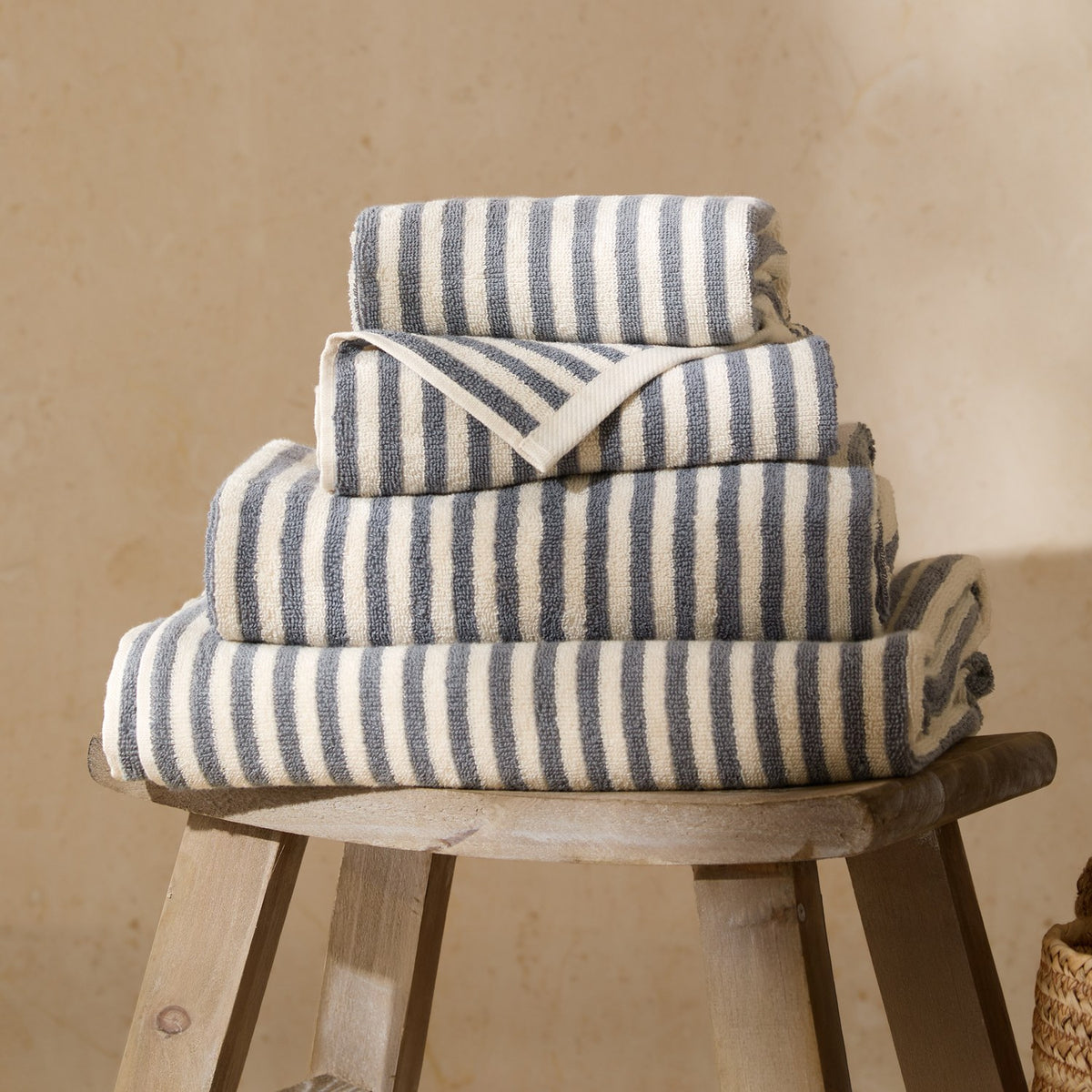 Stacked Hebden Striped Cotton Towels in mineral blue and cream, folded in varying sizes on a wooden stool against a neutral background.