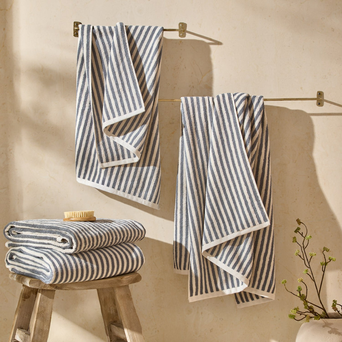 Hebden Striped Cotton Towels in mineral blue and cream, hung on a brass rail and stacked folded on a wooden stool in a bathroom setting.