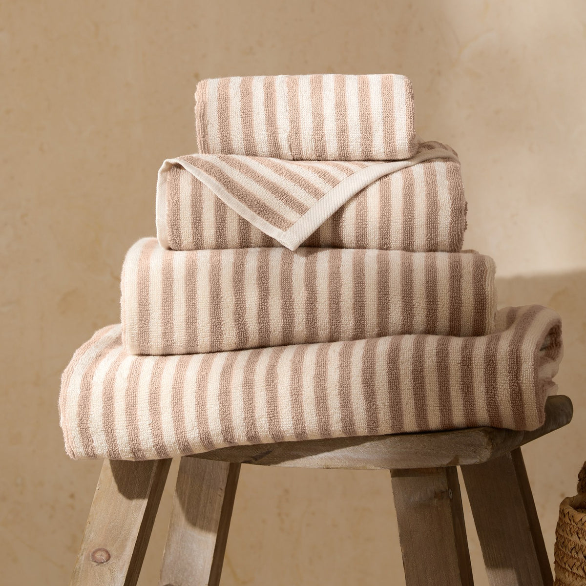 Hebden Striped Cotton Towels in natural beige and cream, stacked in varying sizes with folded edges on a wooden stool against a neutral background.