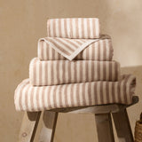 Hebden Striped Cotton Towels in natural beige and cream, stacked in varying sizes with folded edges on a wooden stool against a neutral background.