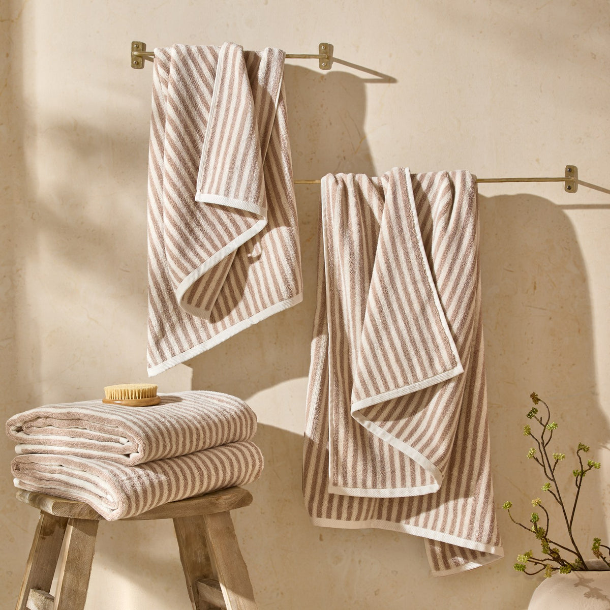 Hebden Striped Cotton Towels in natural beige and cream draped over wall-mounted brass rails and folded on a wooden stool in a bathroom setting.