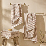Hebden Striped Cotton Towels in natural beige and cream draped over wall-mounted brass rails and folded on a wooden stool in a bathroom setting.