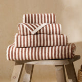 Four folded Hebden striped cotton towels in pecan brown and cream, stacked in graduated sizes on a wooden stool against a neutral background.