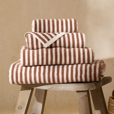 Four folded Hebden striped cotton towels in pecan brown and cream, stacked in graduated sizes on a wooden stool against a neutral background.