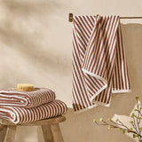 Hebden Striped Cotton Towels in Pecan, folded and hanging in a bathroom setting, showing the reddish-brown and cream vertical stripe pattern on looped cotton terry.