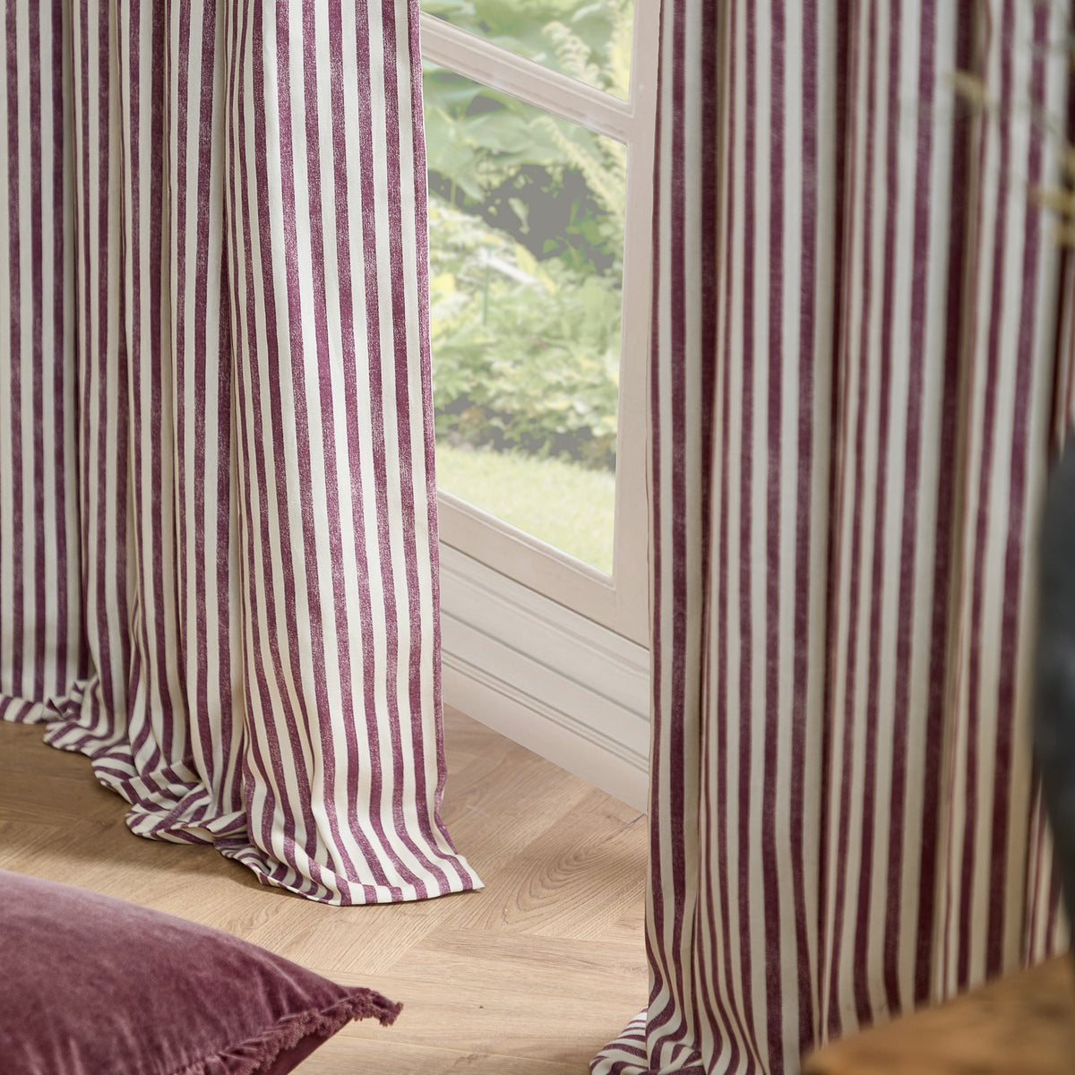 Hebden Cotton Printed Eyelet Curtains in Beetroot, showing beetroot-red and cream vertical stripe fabric hanging beside a window in a living room setting.