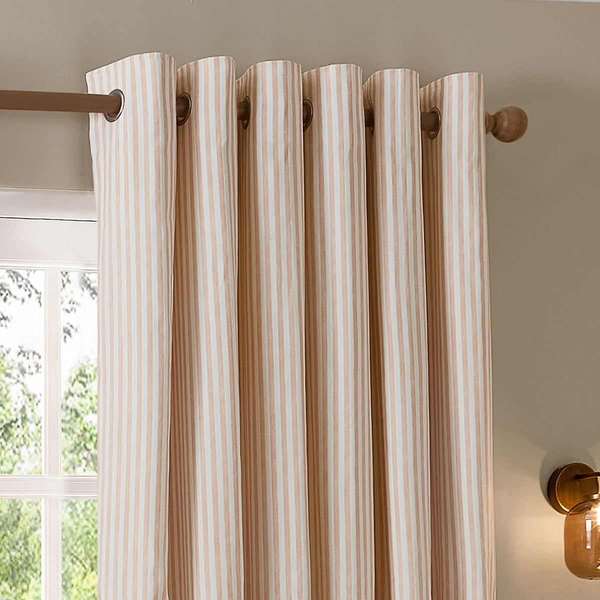 Hebden Cotton Printed Eyelet Curtains in Blush, striped cotton panels with brass eyelets hung on a wooden pole beside a window in a bedroom setting.