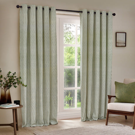 Hebden Cotton Printed Eyelet Curtains in Forest, showing green and cream vertical stripe panels with eyelet header hung on a black rod in a living room window setting.