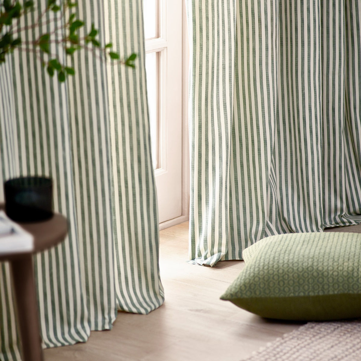 Hebden Cotton Printed Eyelet Curtains in Forest, floor-length cotton panels in a narrow green and off-white stripe hung by a window with a patterned green cushion nearby.