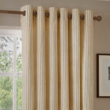 Hebden Cotton Printed Eyelet Curtains in honey-yellow and off-white vertical stripes, shown hanging on a wooden pole beside a window in a living room.
