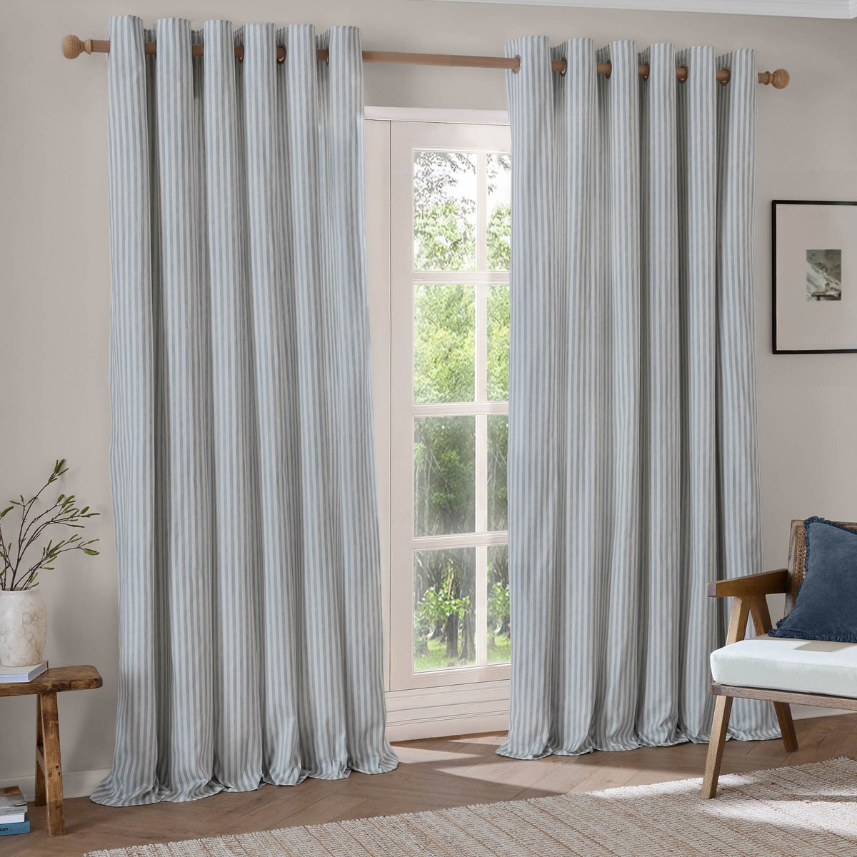 Hebden Cotton Printed Eyelet Curtains in Mineral Blue with vertical stripes, shown as a pair hung on a wooden pole beside a glazed door in a living room.