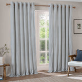 Hebden Cotton Printed Eyelet Curtains in Mineral Blue with vertical stripes, shown as a pair hung on a wooden pole beside a glazed door in a living room.