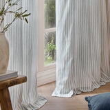 Hebden Cotton Printed Eyelet Curtains in Mineral Blue, striped cotton panels framing a window in a living room with a wooden stool and vase nearby.