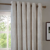 Hebden Cotton Printed Eyelet Curtains in mole brown and cream stripe, hung on a wooden pole beside a window in a living room setting.