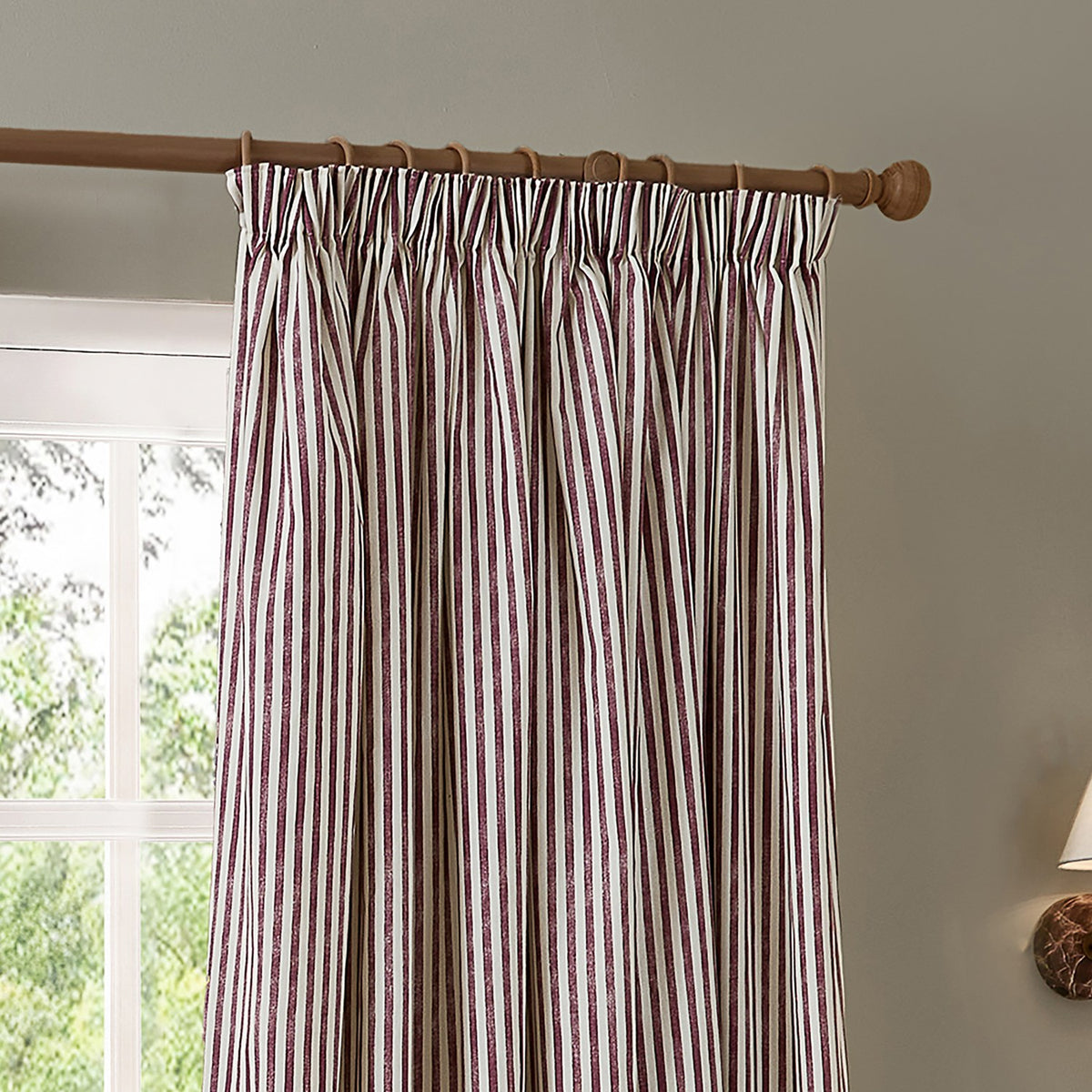 Hebden Striped Cotton Pencil Pleat Curtains in beetroot red and cream, gathered on a wooden pole beside a window in a living space.