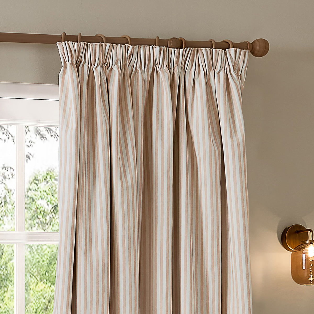 Hebden Striped Cotton Pencil Pleat Curtains in blush and off-white, shown hung on a wooden pole beside a window in a living room setting.