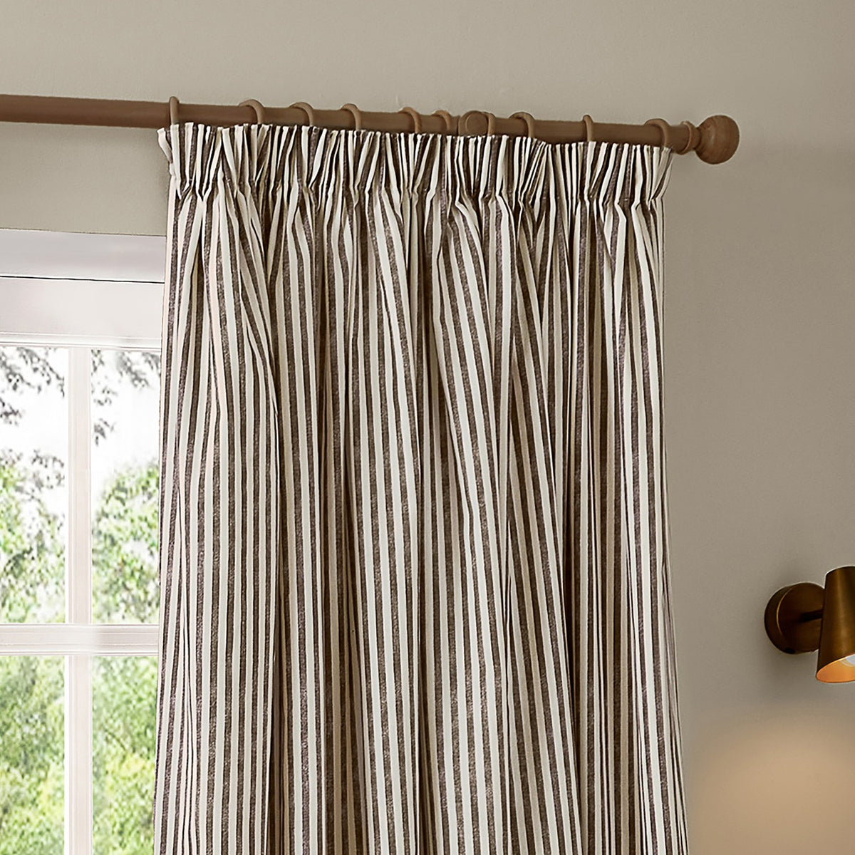 Hebden pencil pleat curtains in chocolate brown vertical stripe on off-white cotton, shown hanging from a wooden pole beside a window in a living room setting.