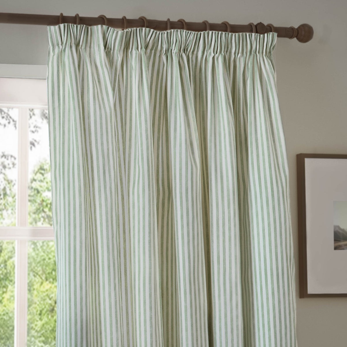 Hebden pencil pleat curtain in eucalyptus green striped cotton, shown hanging on a wooden rod beside a window in a living room setting.