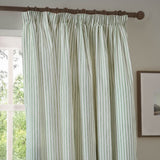 Hebden pencil pleat curtain in eucalyptus green striped cotton, shown hanging on a wooden rod beside a window in a living room setting.