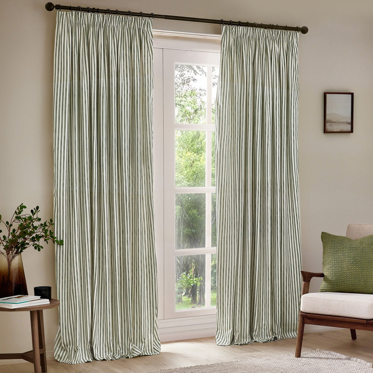 Hebden Striped Cotton Pencil Pleat Curtains in Forest, a pair with green and off-white stripes hung on a dark pole in a living room beside a window.