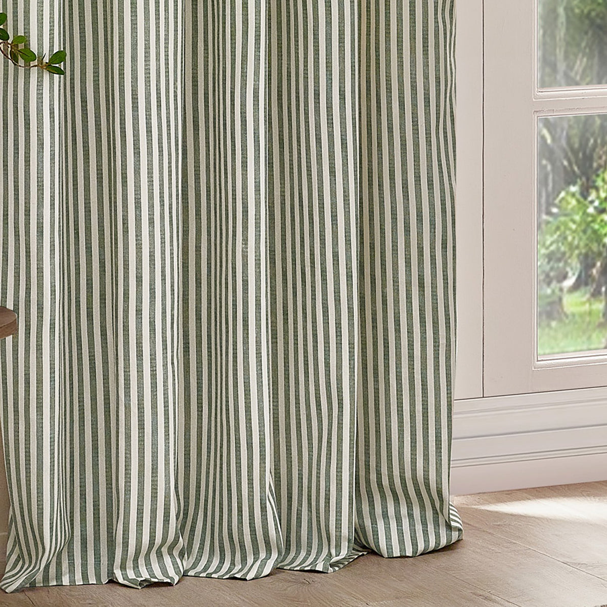 Hebden Striped Cotton Pencil Pleat Curtains in Forest, showing floor-length off-white fabric with narrow green vertical stripes hanging beside a window in a bright room.