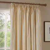 Hebden pencil pleat curtain in honey-yellow and cream cotton stripe, shown pleated on a wooden pole beside a window in a living room setting.
