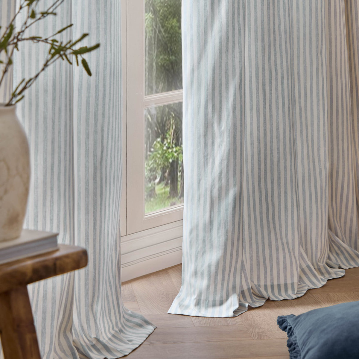 Hebden Striped Cotton Pencil Pleat Curtains in Mineral Blue, floor-length with vertical blue-and-white stripes, shown hanging beside a window in a living room setting.