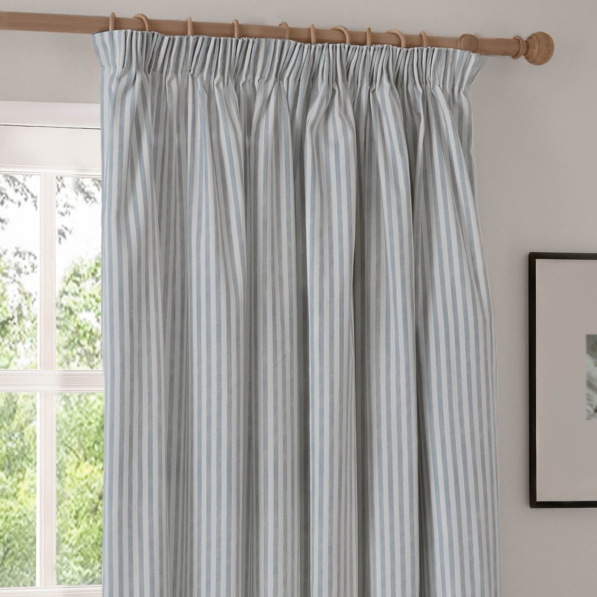 Hebden Striped Cotton Pencil Pleat Curtains in Mineral Blue, showing the blue-and-white vertical stripe and gathered header hung on a wooden pole in a living room.