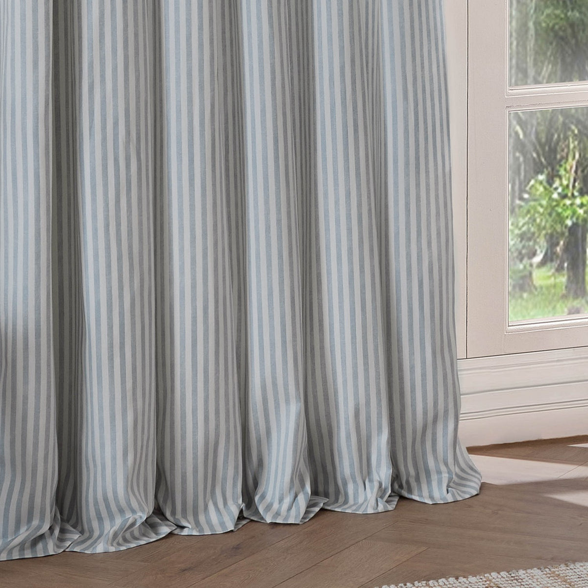 Hebden Striped Cotton Pencil Pleat Curtains in Mineral Blue, showing pale blue and white vertical stripes with a gathered header, styled beside a window in a living room.